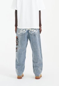 Volné džíny z denimu ve světle modré barvě s vyšitými detaily na levé noze a logem "Ed Hardy" vyšitou na zadní straně. Nosené s bílým tričkem.