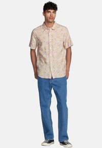 Kortärmad skjorta med knapp i ljusbeige med ett mönster av abstrakta solrosor. Matchad med blå jeans och svarta sneakers.