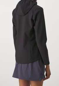 Giacca softshell nera con cappuccio, caratterizzata da una texture liscia e un design aderente, indossata sopra una gonna viola scuro.