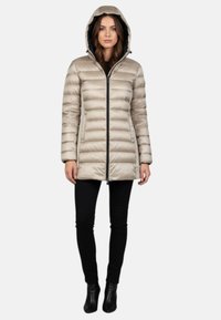 Dames beige pufferjack met capuchon, ritssluiting, horizontale stiksels en lange mouwen, gecombineerd met zwarte nauwsluitende broek en laarzen.