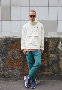 Cremefarvet hættejakke med en frontlomme, teal joggers og navy atletiksko med orange detaljer. Briller på. Tekstureret væg som baggrund.