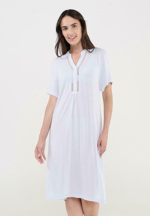 Mujer con un vestido blanco de manga corta, largo hasta la rodilla, con escote en V y tapeta con botones, sonriendo frente a un fondo claro y uniforme.