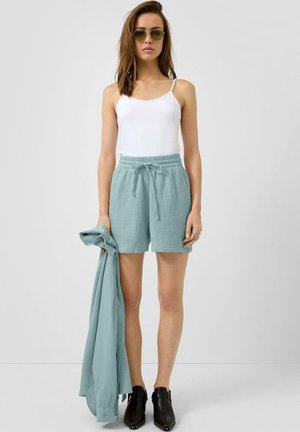 Vrouw draagt groene short met trekkoord, wit mouwloos topje, groene zonnebril, zwarte enkellaarzen, houdt bijpassend groen shirt met lange mouwen vast.