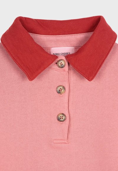 Ljusrosa polo med en kontrasterande röd krage, treknappsstängning och en tygetikett som visar "ROBO CHOSES". Mjuk, texturerad bomullsmaterial.