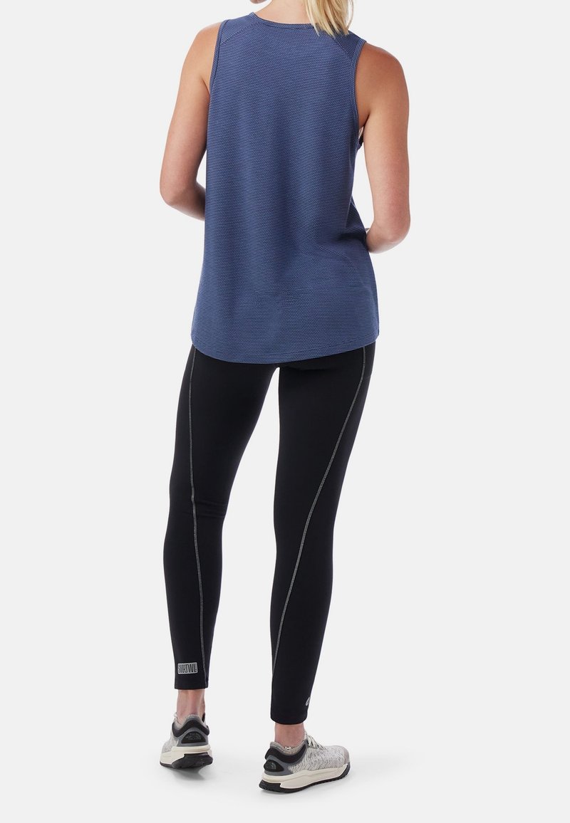 Femme portant un haut bleu sans manches, des leggings noirs avec des coutures et des baskets de course grises, debout, dos tourné, sur un fond blanc.