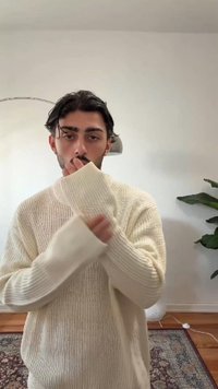 Gestrickter cremefarbener Pullover mit geripptem Muster und übergroßen Ärmeln, mit hohem Kragen und lockerem Sitz. Im Hintergrund ist eine Pflanze zu sehen.