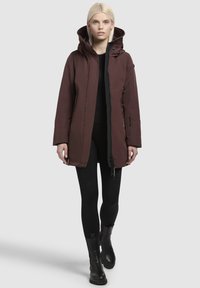 Bordeauxrode parka-jack met een capuchon, rits aan de voorkant, twee zijzakken en een gestructureerde voering. Draagt over een zwarte top en legging, gecombineerd met zwarte laarzen.