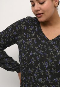 Blouse noire avec un motif floral en violet et vert. Design en V avec des manches longues bouffantes et un tissu texturé.