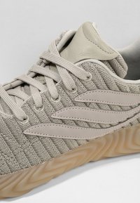 Sneaker med en ljusgrå texturerad stickad ovansida, off-white läderdetaljer och en skulpterad gummisula. Har snören och design med tre ränder.