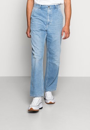 Homme portant un jean droit bleu clair et des baskets blanches à semelles épaisses, debout sur un sol gris devant un mur uni.