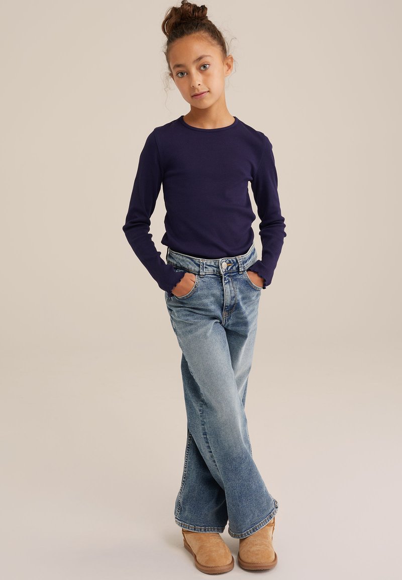 Tengerész kék, hosszú ujjú póló bordás textúrával, világoskék trapéznyakkal és világosbarna csizmával párosítva. Az öltözet klasszikus szabású.