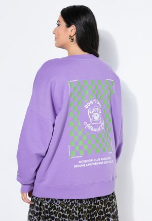 Nainen pukeutunut violettiin svetariin, jossa on vihreä ruutukuvio ja teksti "DON'T CALL Antisocial Club" selässä, yhdistettynä leopardikuvioiseen hameeseen.