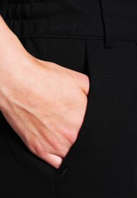 Ruka vložená do přední kapsy černých kalhot s viditelným švem a malou textilní cedulkou poblíž okraje kapsy.