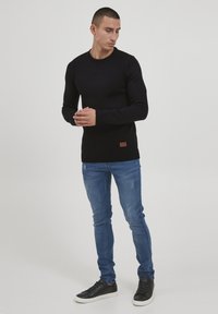 Fekete kötött pulóver bordás mandzsettával és szélekkel, sérült kék farmernadrággal és fehér talpú fekete tornacipővel párosítva.