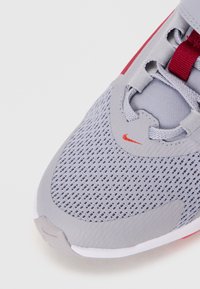 Baskets de sport grises avec un upper en maille, accents rouges et semelle texturée. Le design comprend des lacets avec œillets renforcés et un détail de logo.