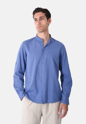 Uomo che indossa una camicia blu con collo alla mandarino abbottonata e pantaloni beige, in piedi con una mano in tasca davanti a uno sfondo uniforme.