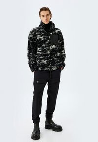 Zwart-wit gemarmerde fleece sweatshirt met een hoge kraag en een zwarte borstzak, gecombineerd met zwarte cargobroek en stevige zwarte laarzen.
