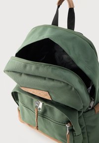 Πράσινο υφασμάτινο σακίδιο με ανοιχτό κύριο διαμέρισμα, μπροστινή τσέπη με φερμουάρ, μαύρες λουρίδες λαβής και καφέ σουέντ διακοσμήσεις σε φερμουάρ και βάση.