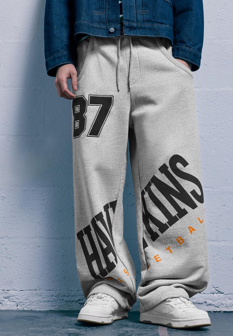 Szürke tréningnadrág, amelyen a "87" és a "HAWKINS BASKETBALL" feliratok nyomtatott grafikával, élénk fekete és narancssárga színekkel jelennek meg. A megjelenést fehér sportcipők egészítik ki.