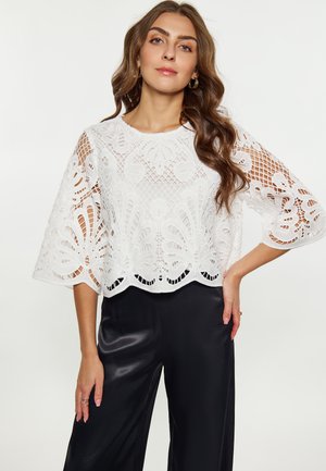 Top corto in pizzo bianco con motivi floreali, dettagli in pizzo e maniche ampie; abbinato a pantaloni neri a vita alta.