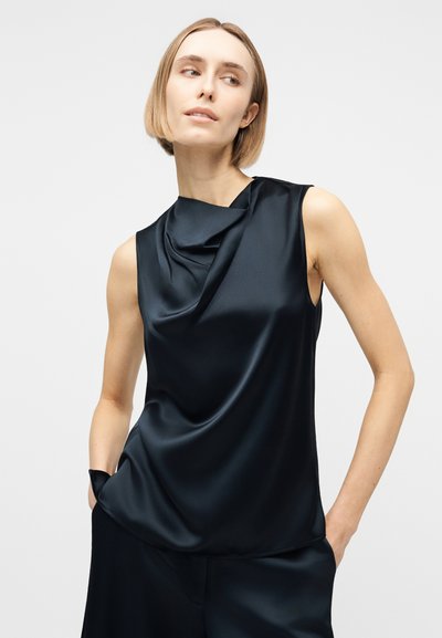 Γυναίκα με κοντά μαλλιά που φοράει μαύρο σατέν τοπ χωρίς μανίκια με ντραπαρισμένο λαιμόκοψη και ταιριαστό μαύρο παντελόνι, με τα χέρια στις τσέπες, κοιτάζει προς τα πλάγια.
