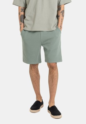 Man wearing sage green textured shorts, light gray t-shirt, black slip-on shoes, standing with hands in pockets, tattooed forearms visible.