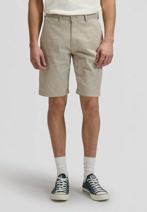 Pantaloncini beige in cotone con taglio dritto, vita standard con chiusura a bottone e risvolti, abbinati a scarpe da ginnastica blu navy.