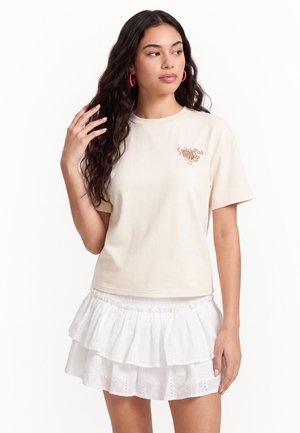 Kvinde med langt, bølget hår iført pink hoop-øreringe, en cremefarvet t-shirt med lille grafik og et hvidt mini-nederdel med lag og huller.