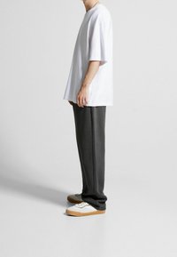 Biała, oversize koszulka z krótkim rękawem, zestawiona z ciemnoszarymi, zwężanymi spodniami dresowymi oraz białymi trampkami z beżowymi akcentami. Widoczne gładkie faktury tkanin.