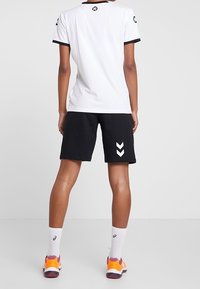 Bílá atletická košile s černými akcenty, v kombinaci s černými šortkami s chevronovým vzorem. Celý vzhled doplňují světlé ponožky a boty.