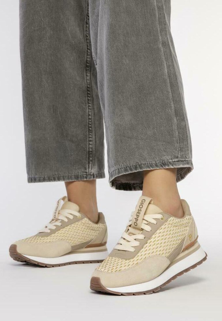 Zapatillas deportivas beige con parte superior de malla, suela de goma y acentos sutiles. Los cordones son claros con un logo en el lateral y detalles texturizados.