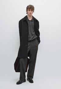 Zwarte overjas, grijs gebreide polo, gestreepte broek en zwarte schoenen. Het onderwerp houdt een kastanjebruine tas vast. Lichte achtergrond versterkt het contrast.