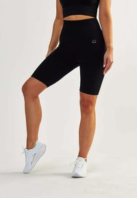 Black high-waisted bike shorts with textured fabric, mid-thigh length, and a small heart logo on the side, paired with white athletic shoes.
