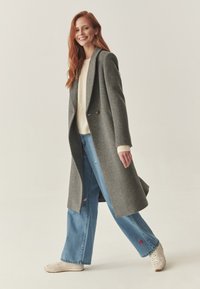 Abrigo de lana gris con un diseño entallado, amplios solapas y dos botones. Combinado con un suéter crema, jeans azules y zapatillas claras.