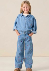Light blue denim shirt and wide-legged jeans with red heart patterns. Shoes are brown with laces. The shirt has a collar and buttons.