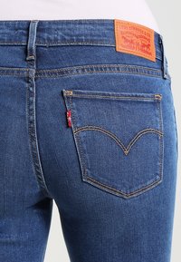 Modré džínové kalhoty s zadní kapsou, která má červenou štítek a oranžové šití. Pas má hnědý kožený štítek s logem značky.