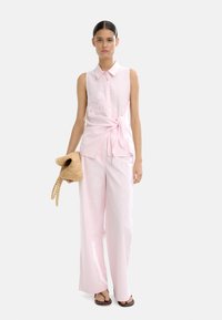 Jasnoróżowa bluzka bez rękawów z wiązaniem z przodu i haftowanymi detalami, w połączeniu z dopasowanymi spodniami o szerokich nogawkach oraz plecioną torbą.