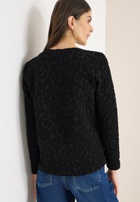 Černý svetr s texturou a vystouplým vzorem leopardí kůže. Má dlouhé rukávy a volný střih, je spárovaný s modrými džínami.