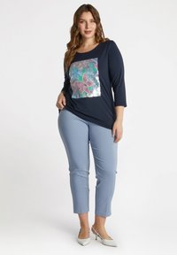 Marineblauwe langemouwtop met een bloemig paillettenontwerp op de voorkant en lichtblauwe fitted broeken. De broeken hebben een cropped lengte en een gladde textuur.