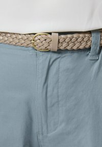 Przeplatany beżowy pasek z okrągłą złotą klamrą, noszony na jasnoniebieskich spodniach z bocznymi kieszeniami i miękką teksturą.