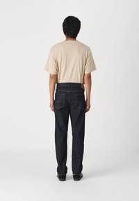 Beige t-shirt, lös passform, korta ärmar; mörkblå raka jeans med framträdande sömmar och två bakfickor. Skor är inte synliga.