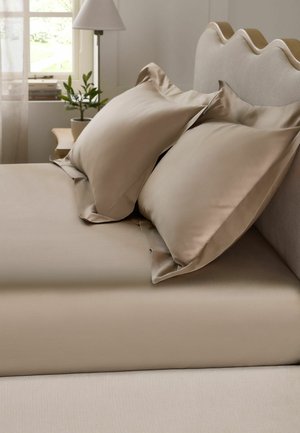 Beiges Bett mit glatten Laken und zwei großen passenden Kissen an einem gewellten Kopfteil, mit einer kleinen Pflanze und einer Lampe im Hintergrund.