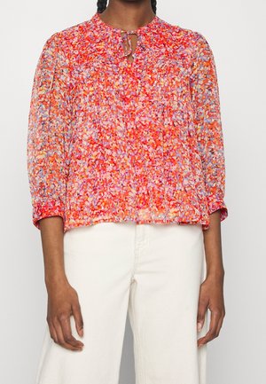 Kvinde iført en livlig rød og orange blomstret bluse med lange ærmer og en bindedetalje ved halsen, kombineret med højtaljede, lysebeige bukser.