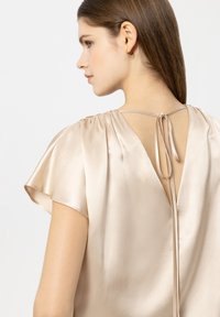 Beige Satinbluse mit tiefem V-Rücken, gerafften Schultern und einem dünnen Bindeband. Glatte Textur mit kurzen Flatterärmeln.