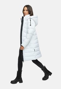 Mujer caminando, vestida con un abrigo largo acolchado blanco con capucha, pantalones ajustados negros y botas gruesas negras sobre un fondo liso.