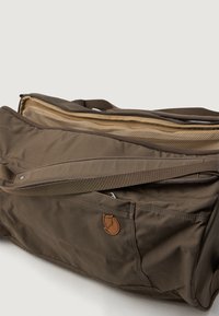Saco de viagem em tecido castanho com uma superfície texturizada, fecho de zíper e um emblema de couro. Inclui um compartimento principal espaçoso e um bolso lateral.