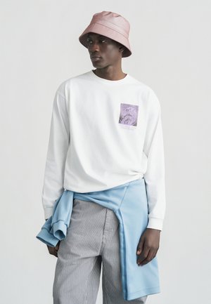 Mladý muž s ružovým klobúkom typu bucket, bielou dlhými rukávmi s grafikou, pruhovanými nohavicami a modrou mikinou zviazanou okolo pása.