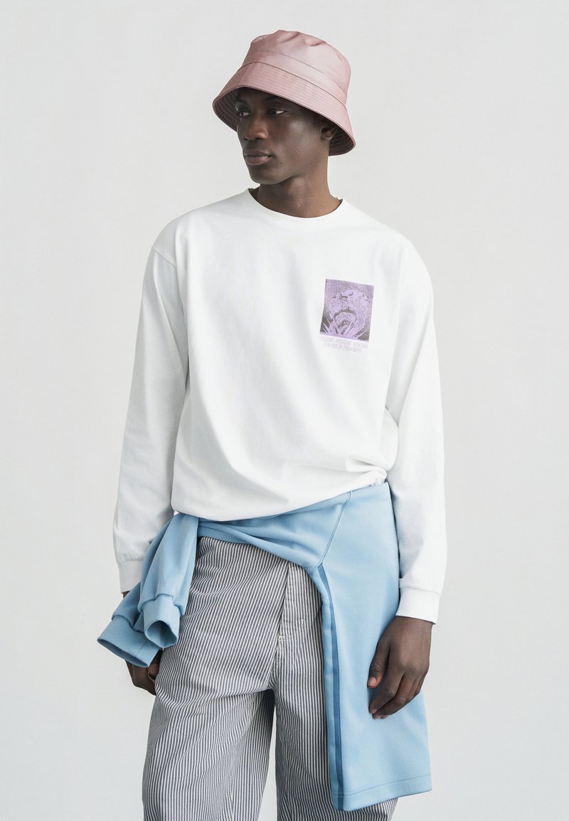 Νεαρός άνδρας με ροζ καπέλο τύπου μπαρμπέρι, λευκό μακρυμάνικο πουκάμισο με γραφικό σχέδιο, ριγέ παντελόνι και μπλε φούτερ δεμένο γύρω από τη μέση του.