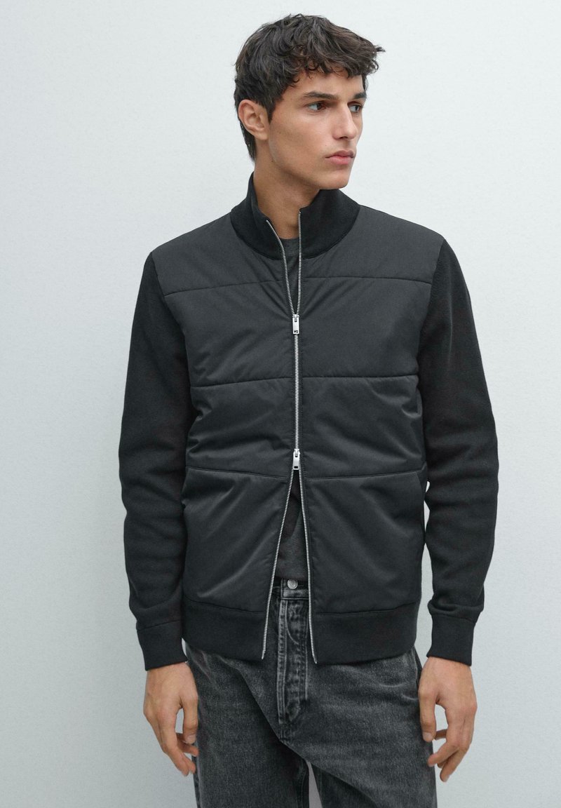 Giacca nera con parte frontale imbottita, colletto alto e chiusura con zip. Maniche lunghe, polsini elasticizzati e vestibilità comoda. Abbinata a jeans scuri in denim.