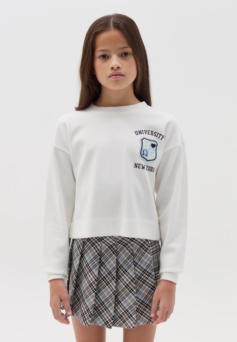 Witte textuurtrui met borduursel "University New York", gecombineerd met een geruite plooiskirt in bruine, blauwe en zwarte tinten.